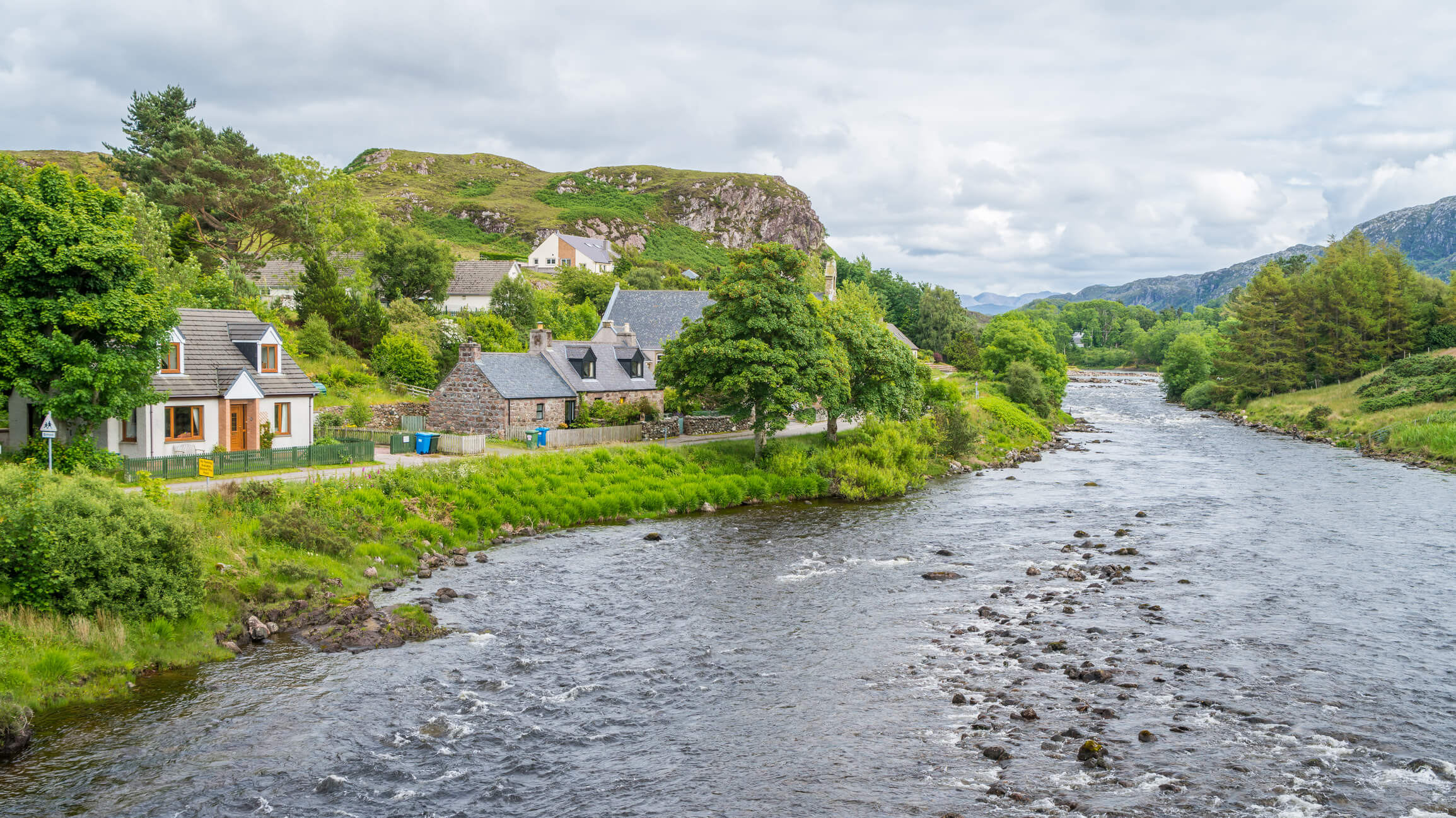 A river flowing through Poolewe with mountains in the distance against a cloudy sky. Houses line the riverside and the sides of the mountains.