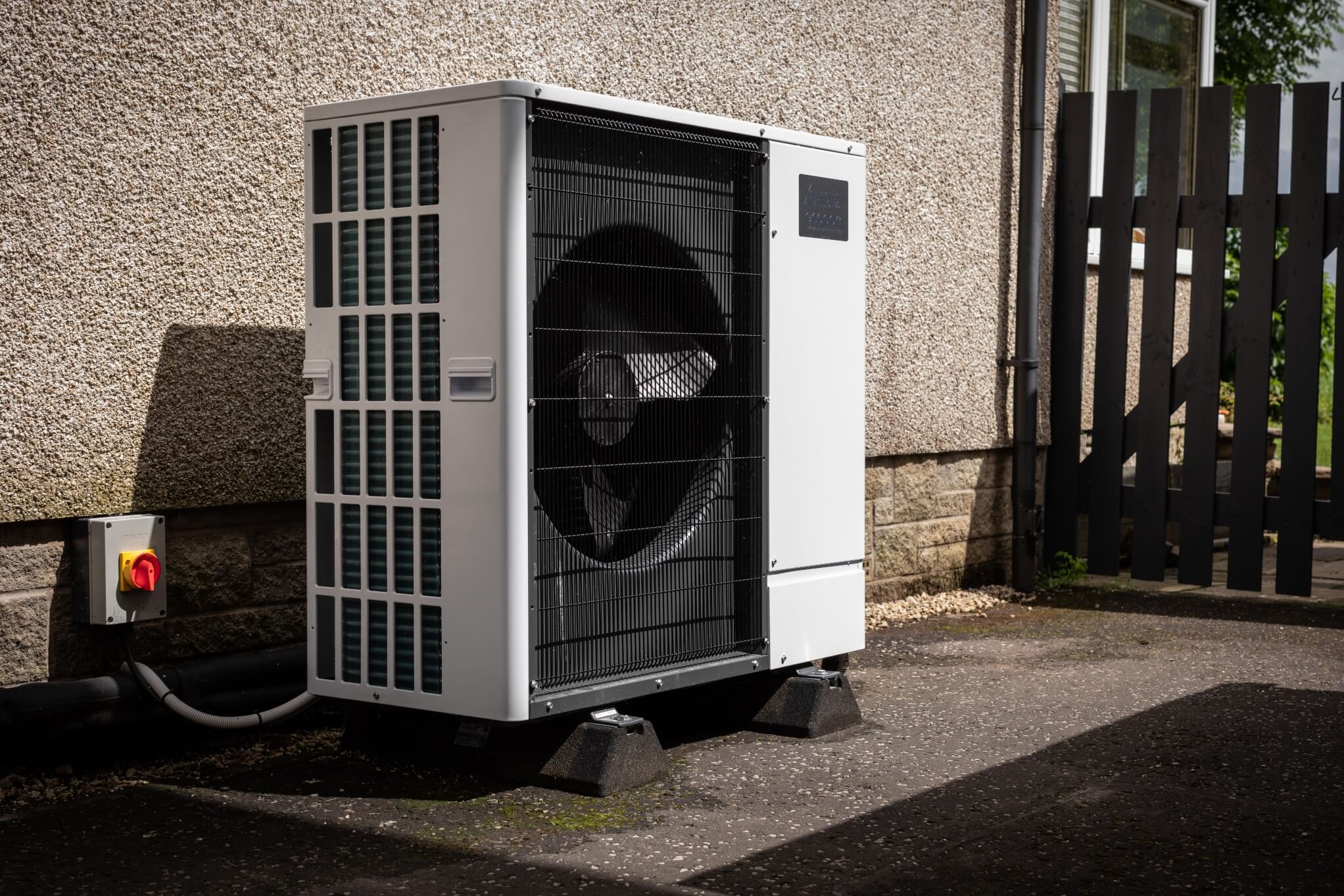 An air source heat pump up against a grey house wall with a small dark brown fence on the right