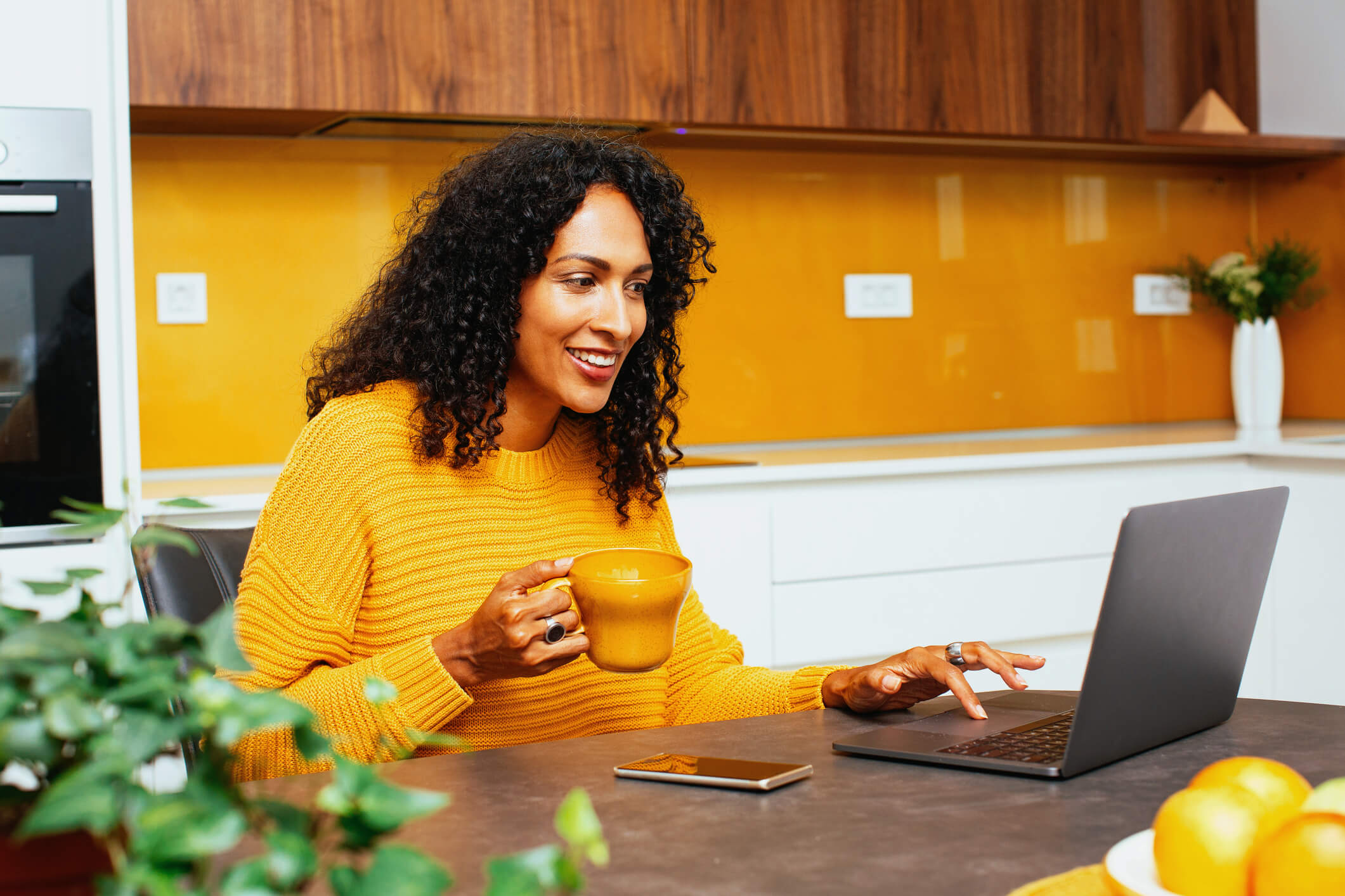 Woman with dark curly hair using computer to see what loans she qualifies for in her stylish kitchen with walls that match her yellow jumper