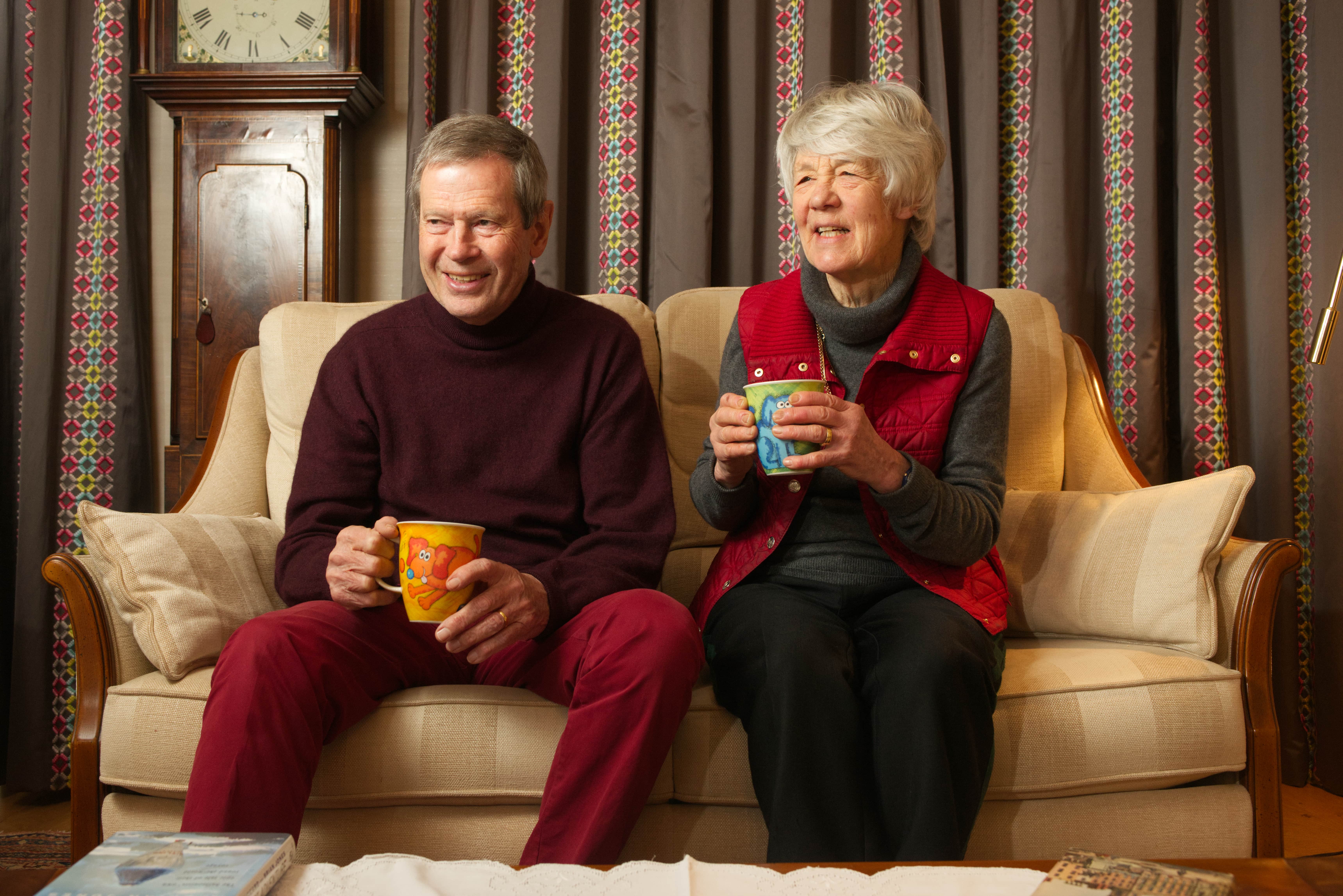 An old couple sitting on their cream sofa next to their grandfather clock. Both of them are holding colourful mugs with cartoon drawings on them