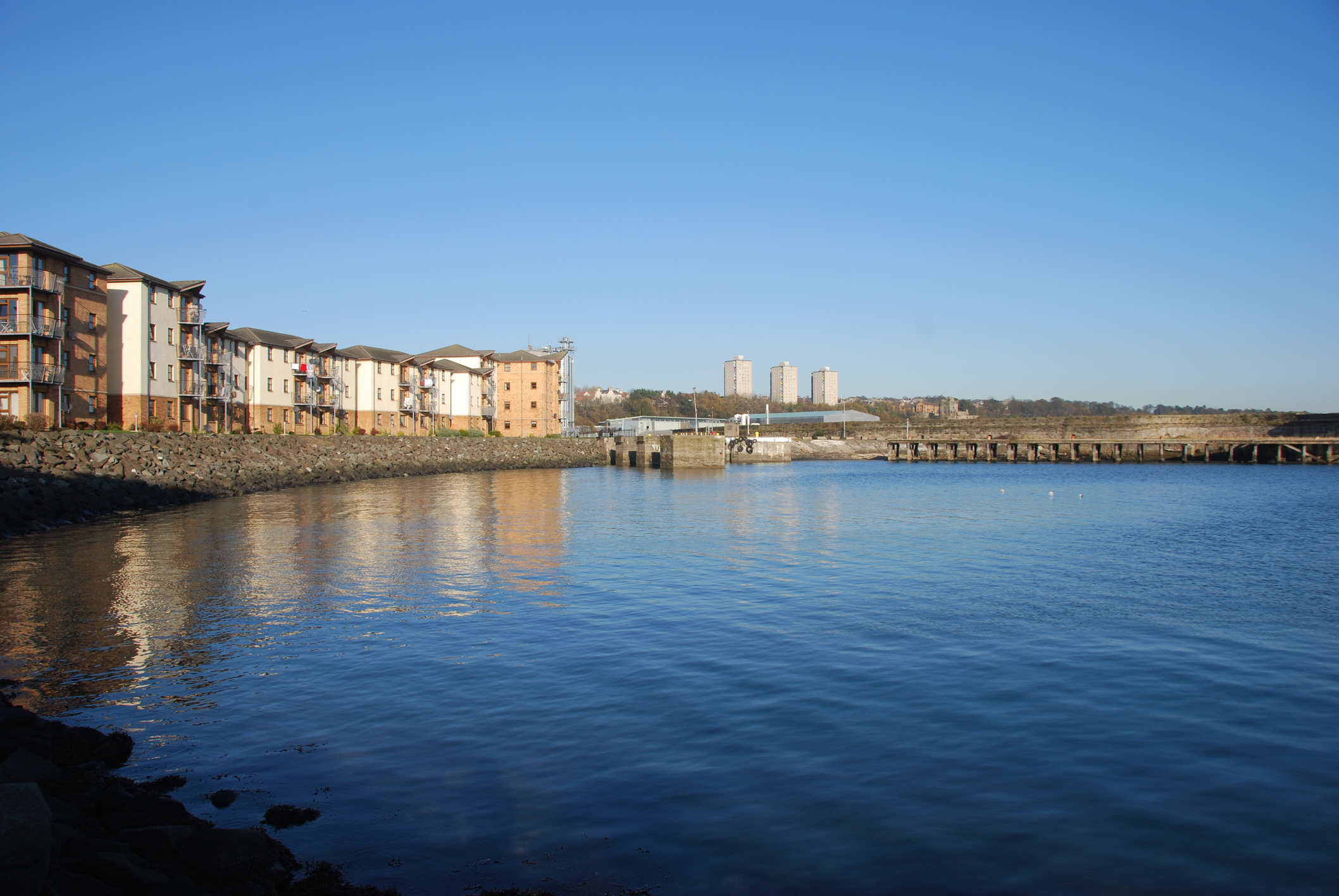 A view of modern apartment buildings at a redeveloped harbour in Kirkcaldy. The water is calm and reflects the clear blue sky.