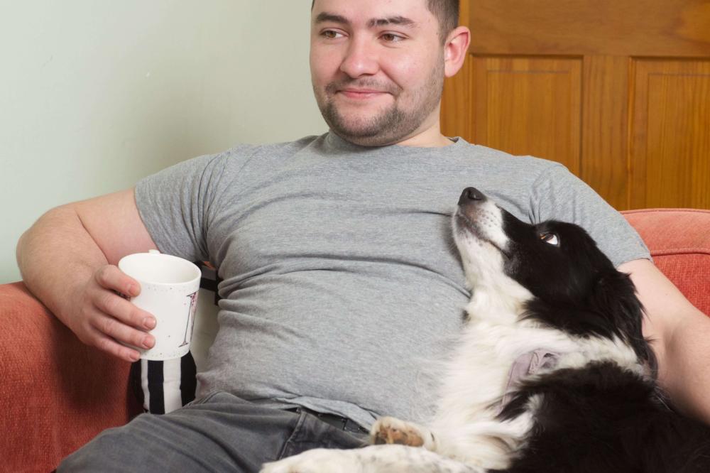 A man sits on his red sofa with a hot drink in a white mug. His black and white dog is sitting next to him and looking at him.